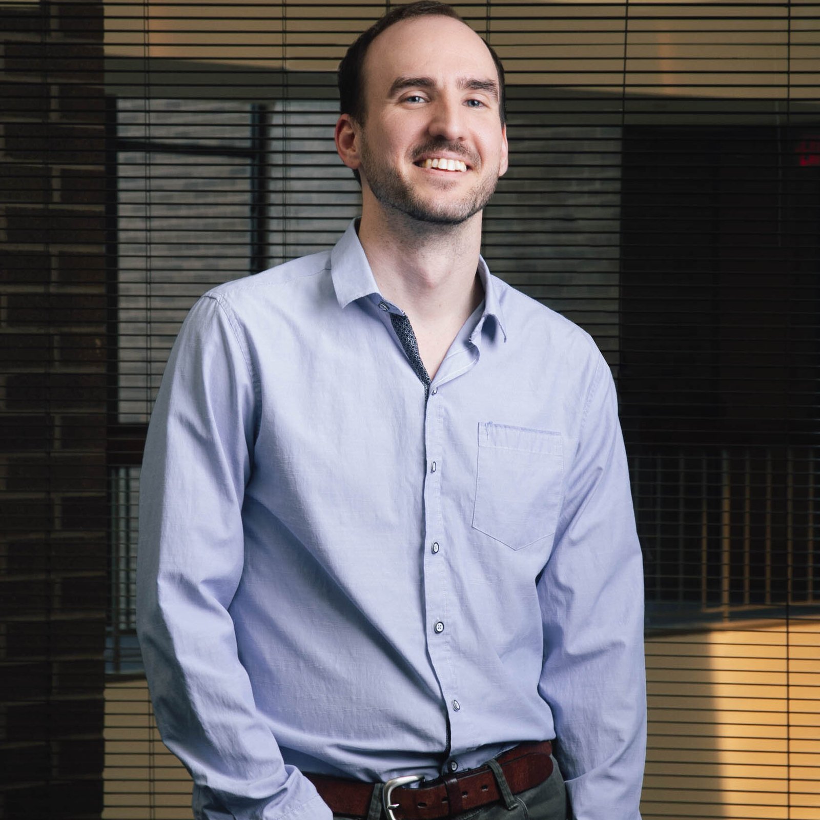 Warm illumination surrounds a smiling man wearing a light blue shirt as he stands indoors in front of horizontal blinds.