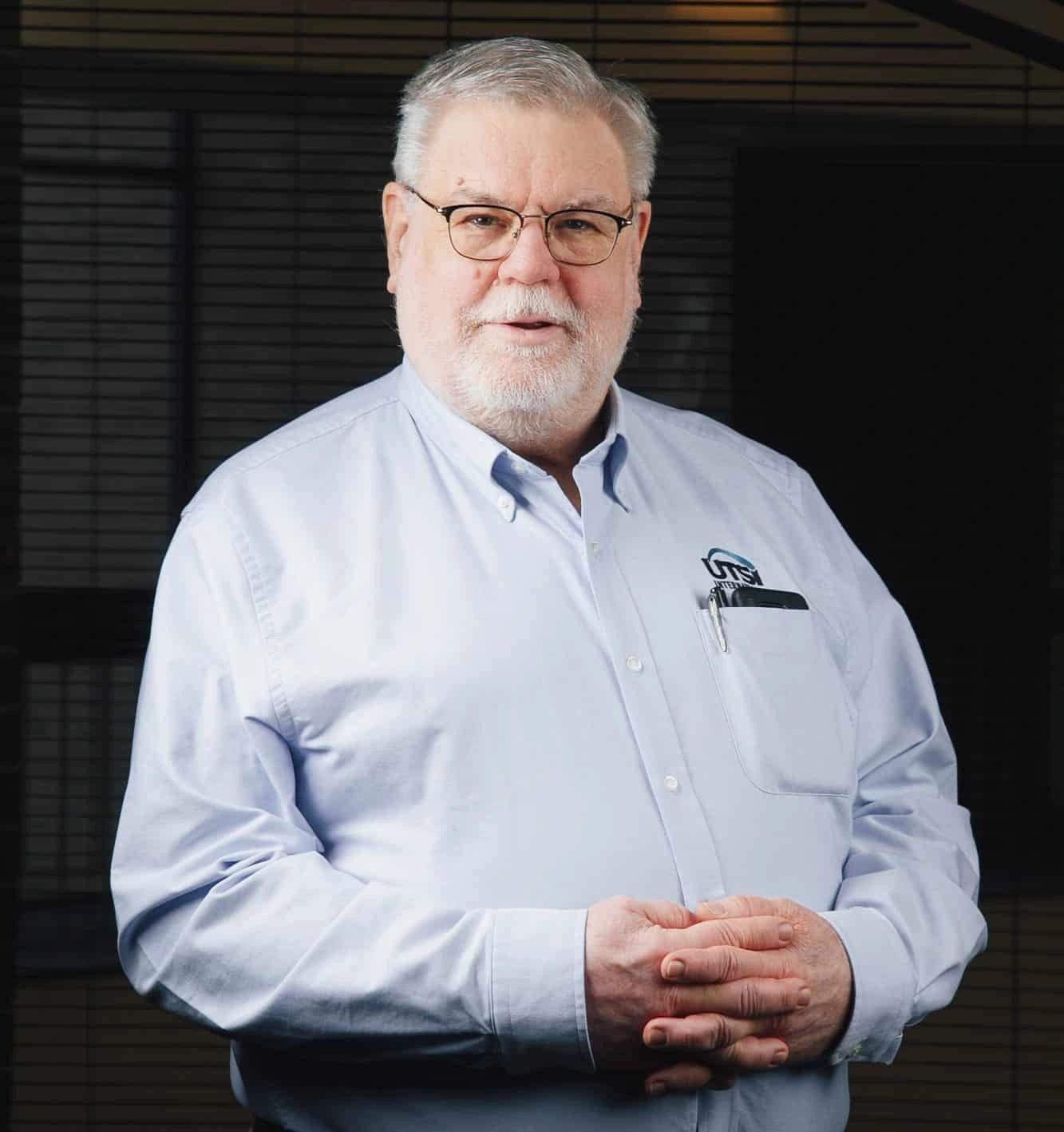 Standing with his hands folded, a man with gray hair, glasses, and a well-groomed beard wearing a light blue shirt exudes confidence.