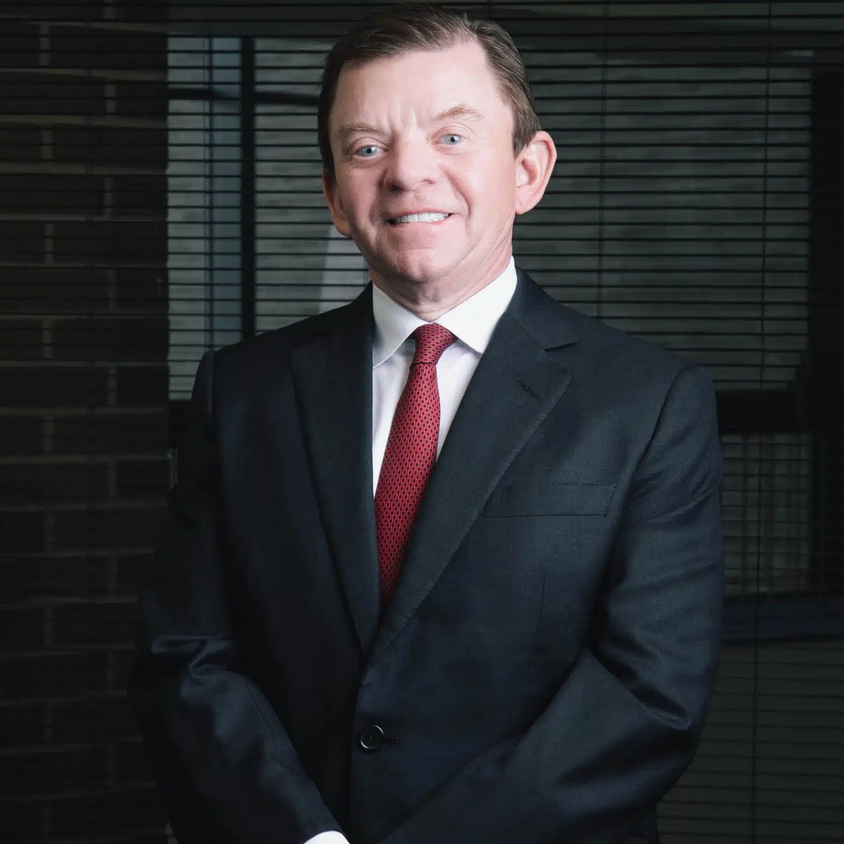 Man in dark suit and red tie smiling, standing with hands folded in front of office blinds in a professional indoor setting.