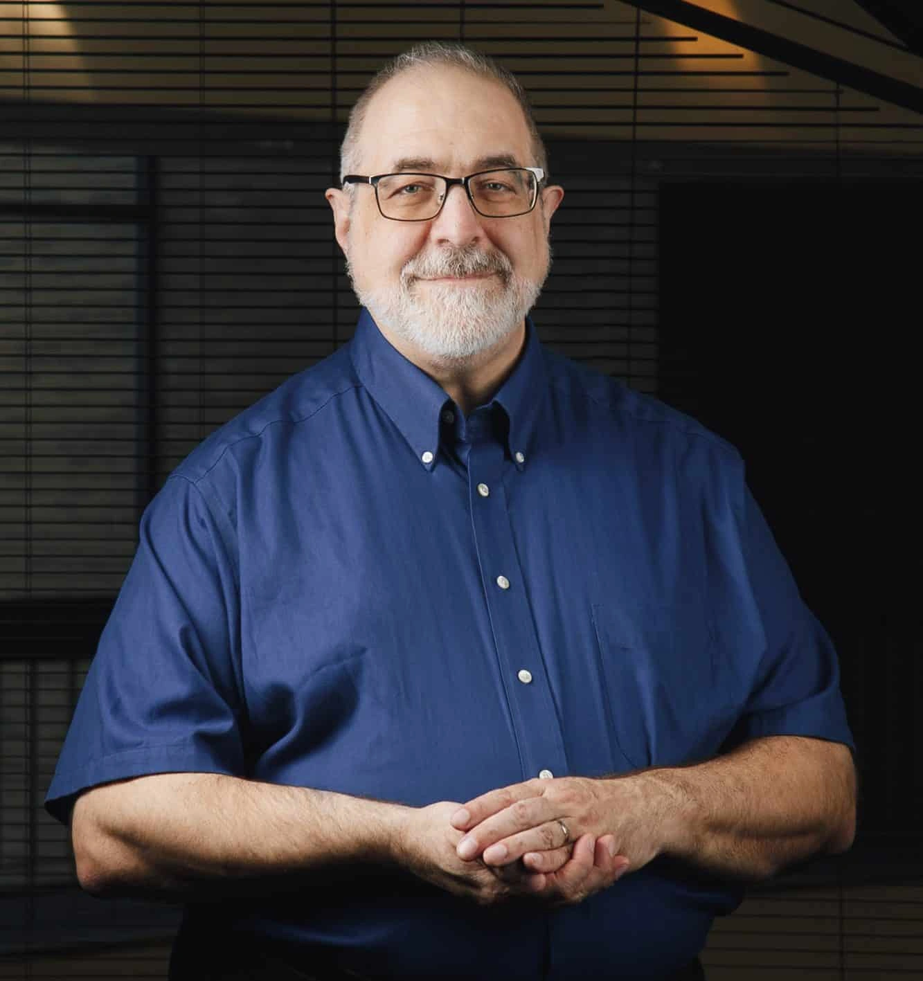 A smiling, white-bearded, glasses-wearing man with a navy blue shirt, his hands clasped gently together, and a confident gaze forward.
