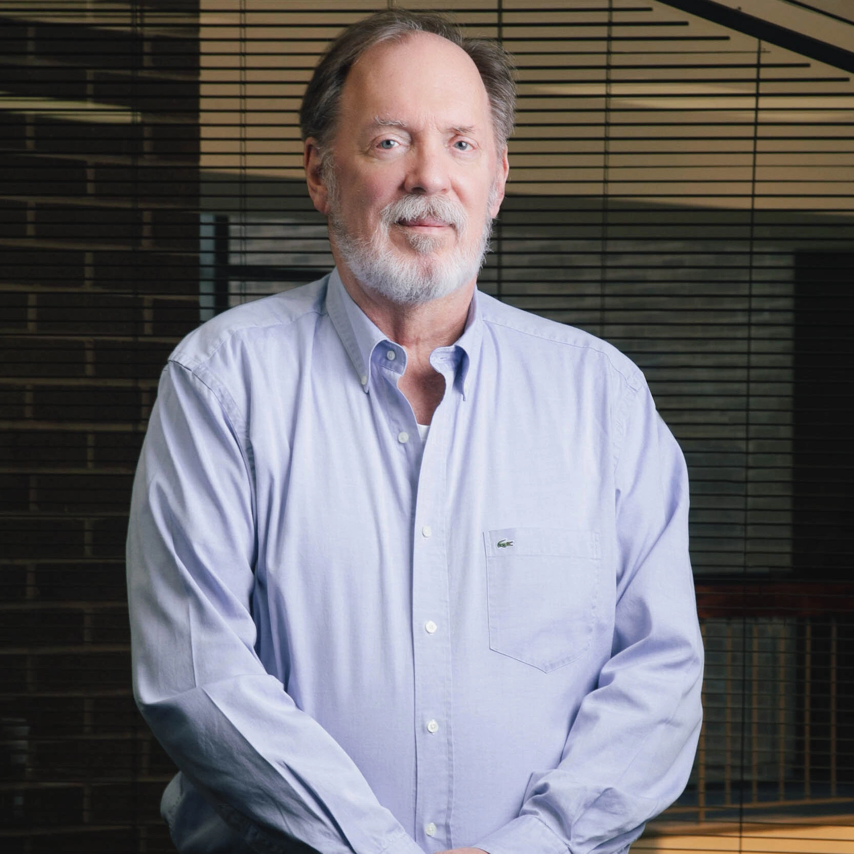 Posing confidently indoors in front of horizontal blinds is an older man with short gray hair and a beard, dressed in a light blue shirt.