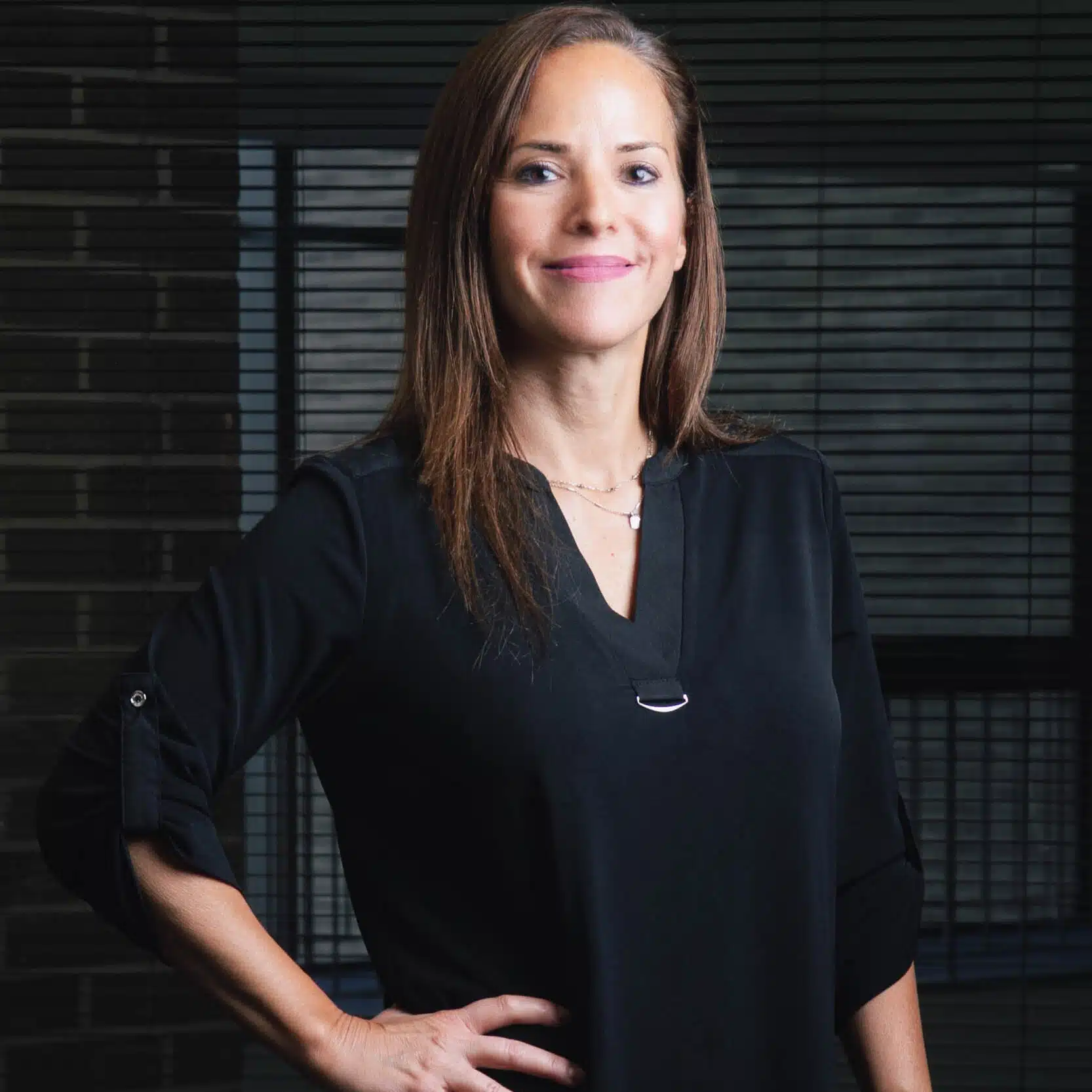 In a professional setting, a self-assured woman wearing a black blouse stands with one hand on her waist and smiles in front of office blinds.